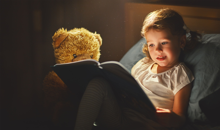 Learning about reading: A little girl sits next to her teddy bear and excitedly reads a book