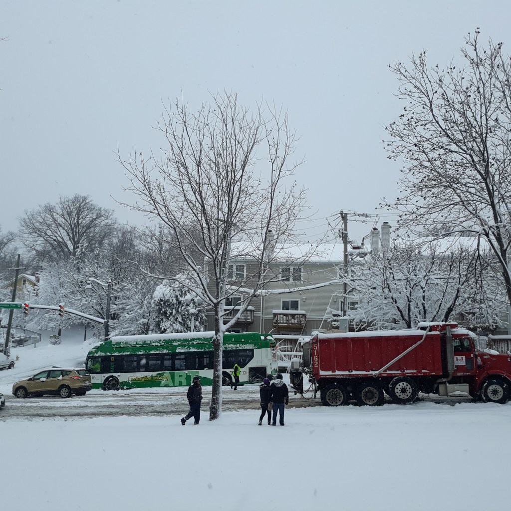On a snowy day, people in black come out to watch a snow plow a bus and an SUV get stuck on the road.